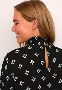 Zwarte blouse met bloemmotief in wit en turquoise, hoge nek met smocking en detail met sleutelgat aan de achterkant, vastgemaakt met twee knopen.
