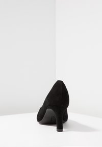 Chaussure à talon haut en daim noir avec un bout pointu et un talon fin, présentant une texture lisse et un logo de marque subtil sur la semelle.