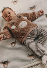 Brun plyschtröja med ko-motiv; randiga byxor; ligger på en filt med ko-mönster; lätt textur. Anmärkningsvärda detaljer inkluderar mjukt tyg.