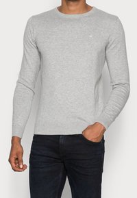 Mann trägt einen hellgrauen Langarmpullover mit gerippten Bündchen und dunkelblaue Jeans, eine Hand in der Tasche, vor einem schlichten Hintergrund.