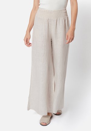 Donna che indossa pantaloni larghi in lino beige chiaro con vita elastica e sandali beige aperti sulle dita, in piedi su sfondo bianco.
