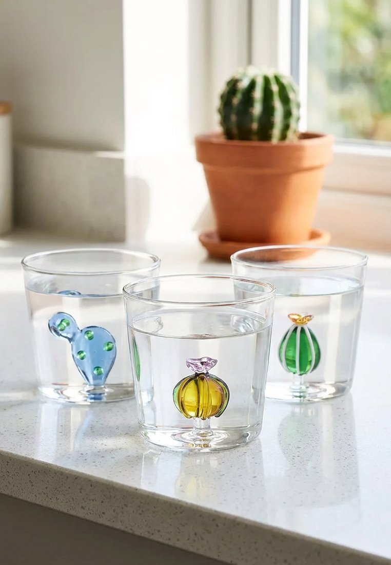 Trzy przezroczyste szklanki napełnione wodą, z każdą zawierającą mały dekoracyjny szklany kaktus, na jasnym blacie kuchennym przy oknie.