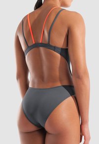 Vue arrière d'une personne portant un bas de maillot de bain gris foncé et un haut assorti avec de fines sangles grises et oranges sur un fond uni.