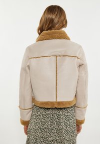 Afskåret læderjakke i lys beige med brunt shearling-krave og kant. Har en glat tekstur og rene sømme, parret med en blomsterkjole.