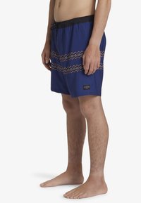 Shorts de bain bleus avec une taille élastique noire, cordon de serrage, et un motif en zigzag rouge et gris à l'avant. Comprend une patte de logo.