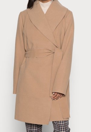 Femme portant un manteau beige ceinturé par-dessus un col roulé blanc et un pantalon à carreaux, debout devant un fond uni.