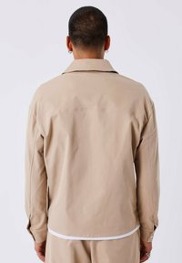 Camicia beige a maniche lunghe con una vestibilità morbida. Presenta un colletto a punta, polsini con bottone e un tessuto liscio e leggero. Vista posteriore.