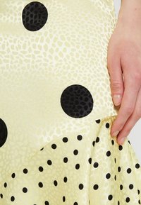 Käsi pitää keltaista kangasta, jossa on suuria ja pieniä mustia pilkkukuvioita sekä hienovarainen tekstuuri.