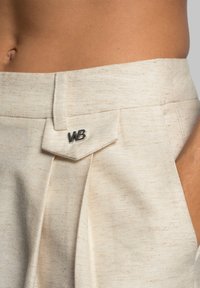 Beige pleated shorts feature a textured fabric with flecks, silver "WB" logo on the waistband, and a tailored silhouette.