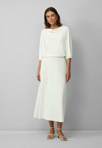Robe longue blanche avec un décolleté drapé et des manches de trois-quarts. Tissu lisse, silhouette semi-ajustée, accompagnée de talons à lanières.