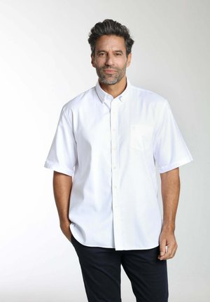 Homme aux cheveux foncés et à la barbe, portant une chemise blanche à manches courtes boutonnée et un pantalon noir, debout avec une main dans sa poche.