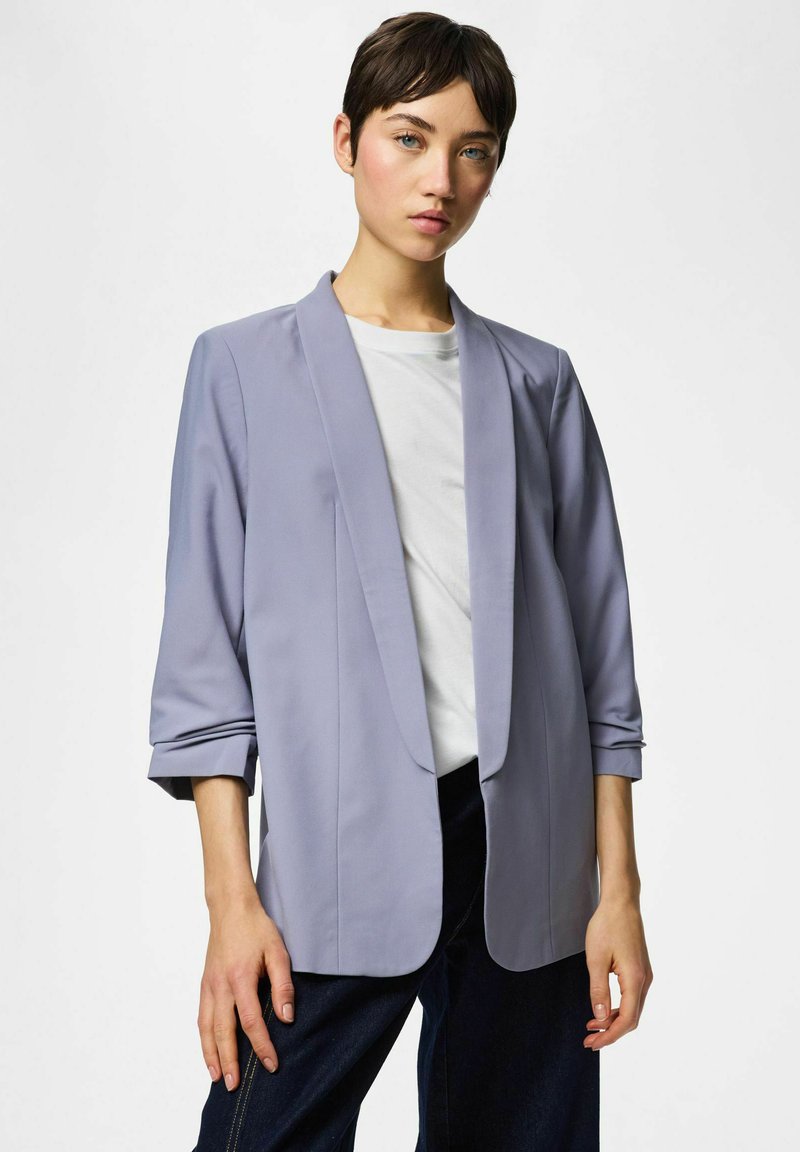 Jeune personne aux cheveux courts et foncés portant un blazer violet clair sur une chemise blanche et un pantalon foncé, debout devant un fond uni.