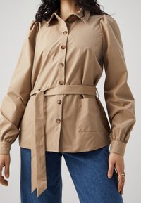 Modellen bär en ljusbeige knapp-up skjorta med knytskärp i midjan och puffärmar, kombinerat med blå jeans, mot en neutral bakgrund.