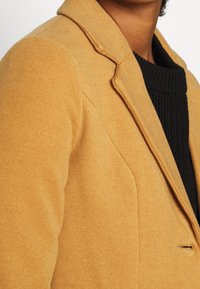 Mustard yellow blazer with a textured fabric, featuring a notched lapel, single front button, and side pockets, layered over a black knit sweater.
