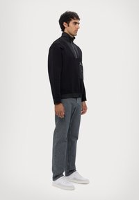 Felpa nera con zip e tasca frontale, abbinata a jeans in denim grigi e sneakers bianche. Texture liscia, vestibilità comoda.