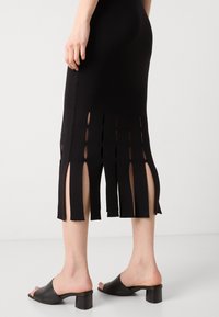 Melnā adīta midi kleita ar sānu šķēlumiem un ovālas formas izgriezumiem. Apvienota ar melnām atvērtām mūlēm ar bloka papēžiem. Gludas faktūras visā.