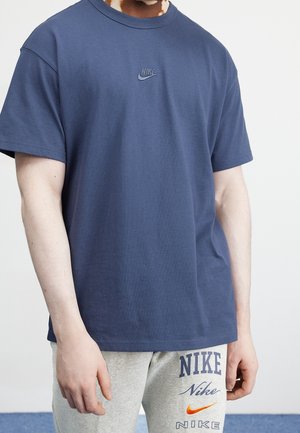 Mand iført en navyblå Nike t-shirt og lysegrå Nike joggingbukser med flere synlige Nike-logoer.