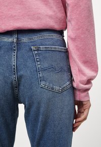 Pantalones vaqueros de mezclilla azul con un corte clásico, que presentan costuras en el bolsillo trasero y una cinturilla alta, combinados con una camisa rosa de manga larga.