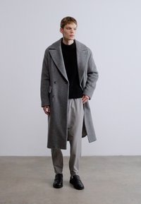 Szürke gyapjúkabát nagy gallérokkal, kétsoros kivitelben, fekete bordázott pulóver és világosszürke nadrág fölött viselve, fekete cipőkkel párosítva.