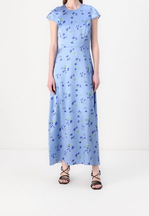 Žena v dlouhých světlemodrých květovaných šatech s krátkými rukávy a černých sandálech na pásky, stojící před bílým pozadím.