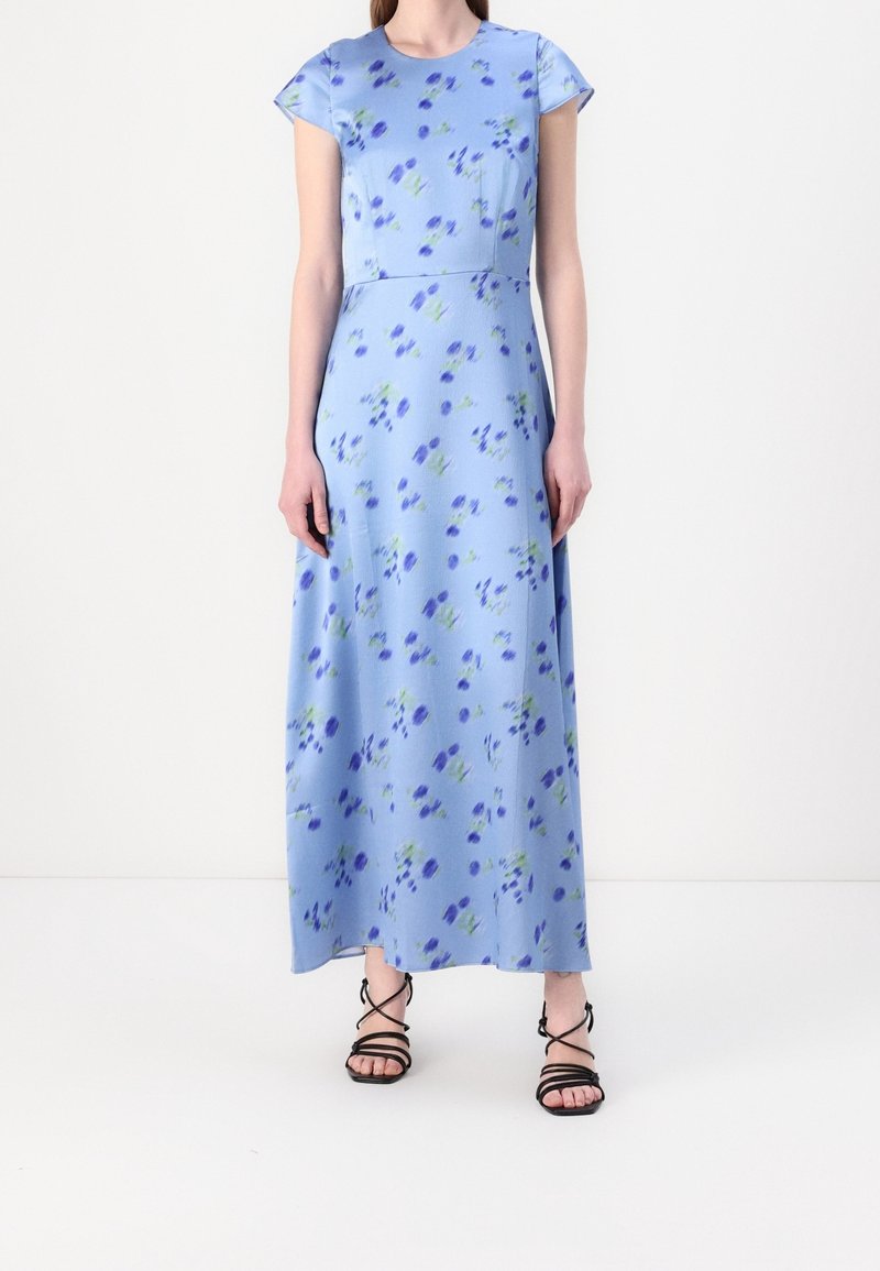 Femme portant une robe maxi florale bleu clair à manches courtes et des sandales noires à brides, debout devant un fond blanc.
