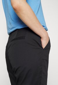 Person iført sorte bukser og en blå T-shirt med korte ærmer, med den ene hånd afslappet placeret i forlommerne på bukserne.