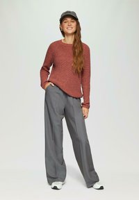 Rostfarbener gestrickter Pullover mit langen Ärmeln, kombiniert mit grauen weit geschnittenen Hosen und weißen Sneakers. Das Model trägt eine graue Kappe.