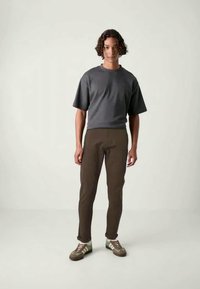 Gray short-sleeve shirt paired with brown fitted pants. The model wears green and beige sneakers, standing against a neutral backdrop.