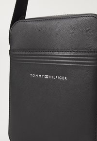 Černá syntetická crossbody taška s texturovaným povrchem. Obsahuje zipové uzavření a embosované logo "TOMMY HILFIGER" na přední straně.