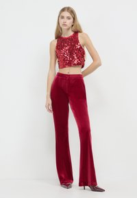 Donna che indossa un top corto rosso con paillettes, pantaloni a vita alta in velluto rosso a gamba larga e scarpe con tacco a punta su uno sfondo bianco semplice.