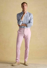 Lichtblauw-wit gestreept overhemd met knoopsluiting, gecombineerd met roze broek met rechte pijpen en beige instapschoenen, tegen een textuurachtergrond.