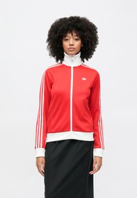 Sieviete ar cirtainiem matiem, tērpusies sarkanā un baltā Adidas sporta jaciņā un melnā svārkā, stāv pret vienmērīgu gaišu fonu.