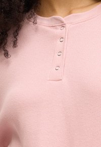 Ροζ τοπ σε ύφανση βάφλ, με στρογγυλή λαιμόκοψη, διακοσμητικά κουμπιά και υφή υφάσματος, που αναδεικνύει έναν απλό και καθημερινό σχεδιασμό.