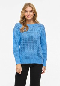 Femme portant un pull en maille légère bleu clair à manches longues et un pantalon noir, debout avec un léger sourire devant un fond blanc uni.