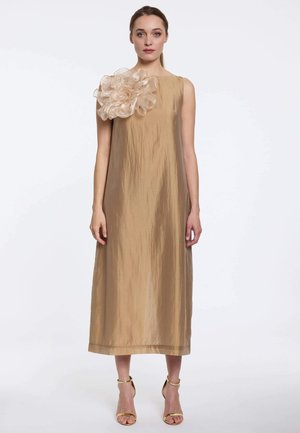 Femme portant une robe sans manches, dorée, longue jusqu’aux chevilles, avec un grand détail floral transparent sur l’épaule gauche et des talons dorés à brides.