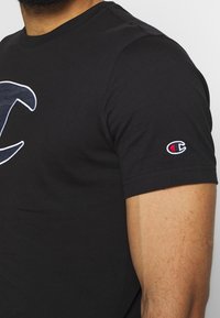 T-shirt in cotone nero con un logo bianco e blu in evidenza sul davanti e un piccolo logo ricamato sulla manica.