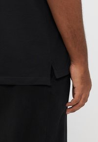 Čierne tričko s bočným rozparkom, vyrobené z textúrovaného materiálu a s bežným strihom. Pohľad zobrazuje lem a časť rukáva.