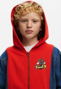 Red zip-up hoodie with a denim left sleeve, featuring a cartoon graphic on the chest, and a soft textured interior.