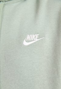 Tejido de color verde claro con un pequeño logo bordado de Nike en blanco, con el swoosh en la zona del pecho de una prenda.