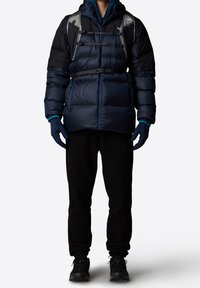 Veste matelassée bleu marine avec capuche, présentant des coutures horizontales, des sangles ajustables et une doublure légère. Associée à un pantalon noir et des gants.