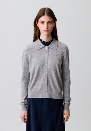 Mujer con pelo largo y castaño lleva una chaqueta de punto ligera gris claro abotonada sobre un vestido azul oscuro con escote fruncido, de pie frente a un fondo liso.