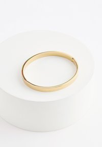 Gold-colored metal bangle bracelet with a smooth finish placed on a white circular pedestal against a plain white background.