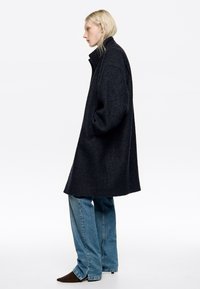 Tumši pelēks mētelis ar augstu apkakli, pārspīlēta silueta, teksturēta auduma, un sānu kabatām, apvienots ar gaiši zilām džinsu biksēm un iegareniem brūniem apaviem.