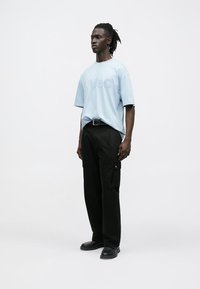 Φαρδιά γαλάζια μπλούζα με ανάγλυφο λογότυπο «HUGO», συνδυασμένη με μαύρο cargo παντελόνι και μαύρα παπούτσια. Απλός, μοντέρνος σχεδιασμός.