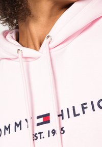 Felpa rosa con occhielli argentati e cordino bianco. Presenta il testo e il logo "TOMMY HILFIGER" ricamati in blu, con la data di fondazione.