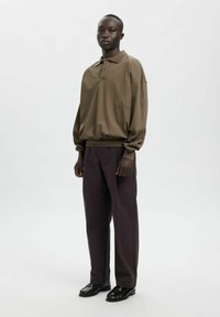 Brūns pārliels polo krekls ar nolocītām plecām, kopā ar tumši violetiem platajiem biksēm un melniem apaviem, uz balta fona.