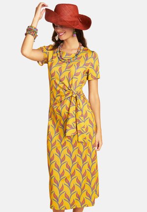 Woman wearing a yellow dress with purple and orange leaf pattern, a red wide-brimmed hat, colorful beaded jewelry, and smiling with eyes closed.