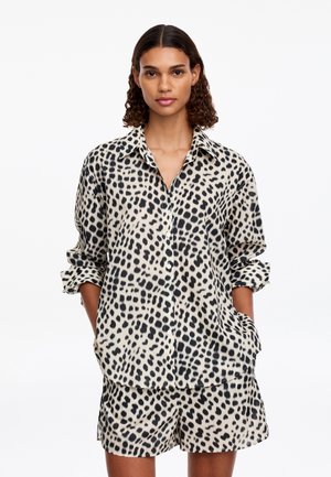 Femme aux cheveux bouclés portant une chemise beige à pois noirs boutonnée et un short assorti, debout les mains dans les poches sur fond blanc.