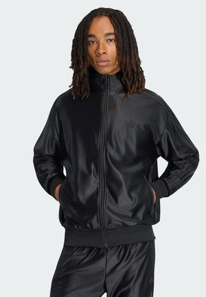 Osoba z dredami do ramion, ubrana w czarną sportową kurtkę na zamek firmy Adidas oraz pasujące spodnie, z rękami w kieszeniach.