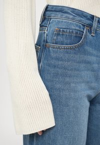Close-up de uma pessoa a usar jeans de ganga azul claro e uma manga de suéter canelado na cor creme a cobrir parte da mão.
